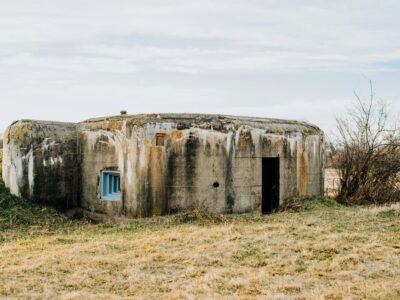Wie viele Bunker hat Albanien? Faszinierende Fakten über das Land der Betonbunker Wie viele Bunker hat Albanien beschäftigt viele Reisende und Geschichtsinteressierte gleichermaßen. Schätzungen zufolge entstanden während der kommunistischen Herrschaft unter Enver Hoxha rund 170.000 bis über 200.000 Bunker, die bis heute das Landschaftsbild des Landes prägen. Von kleinen Betonkuppeln bis zu riesigen unterirdischen Anlagen erzählen die Bauwerke ein außergewöhnliches Kapitel der albanischen Geschichte. Warum so viele Bunker in Albanien errichtet wurden, welche Bedeutung sie heute haben und welche Anlagen besichtigt werden können, erfahren Sie in diesem Artikel. Wie viele Bunker hat Albanien? Schätzungen gehen davon aus, dass es in Albanien zwischen 170.000 und über 200.000 Bunker gibt. Genaue Zahlen existieren nicht, da viele Anlagen nie vollständig erfasst wurden oder im Laufe der Jahrzehnte zerstört beziehungsweise überbaut wurden. Fest steht jedoch, dass kaum ein anderes Land eine vergleichbare Dichte an Verteidigungsanlagen aufweist. Der überwiegende Teil der Bunker in Albanien entstand während der Herrschaft des kommunistischen Diktators Enver Hoxha zwischen den 1960er und 1980er Jahren. Kleine Betonbunker finden sich bis heute entlang der Küste, in den Bergen, auf Feldern und am Stadtrand. Viele stehen noch immer an ihrem ursprünglichen Standort und erinnern an eine Zeit, in der das Land aus Angst vor einer möglichen Invasion auf den Bau hunderttausender Schutzanlagen setzte. Warum hat Albanien so viele Bunker? Der Bau der zahlreichen Bunker in Albanien geht auf die Herrschaft des kommunistischen Diktators Enver Hoxha zurück. Während des Kalten Krieges verfolgte die Regierung eine Politik der Isolation und rechnete jederzeit mit einem möglichen Angriff durch ausländische Staaten. Aus dieser Paranoia heraus entstand eines der größten Bunkerbauprogramme Europas. Zu den wichtigsten Gründen für den massenhaften Bau gehörten: Angst vor einer möglichen Invasion aus dem Ausland. Abschottung Albaniens nach dem Bruch mit dem Warschauer Pakt. Wunsch, die gesamte Bevölkerung im Ernstfall verteidigen zu können. Errichtung kleiner Betonbunker in nahezu allen Landesteilen. Demonstration militärischer Stärke unter der Herrschaft von Enver Hoxha. Vorbereitung auf einen möglichen Angriff, obwohl es dafür keine konkreten Hinweise gab. Viele der Bauwerke wurden jedoch nie militärisch genutzt. Heute gelten sie als sichtbares Symbol für die kommunistische Vergangenheit Albaniens und erinnern an eine Epoche, die von politischer Kontrolle, Isolation und der ständigen Angst vor einer Bedrohung geprägt war. Bau der Bunker unter Enver Hoxha Der außergewöhnliche Umfang des Bunkerbaus lässt sich nur vor dem politischen Hintergrund der damaligen Zeit verstehen. Unter der Herrschaft von Enver Hoxha wurde der Ausbau der Verteidigungsanlagen zu einem zentralen Staatsprojekt, das das gesamte Land über viele Jahre hinweg prägte. Angst vor einer Invasion als Auslöser Unter Enver Hoxha herrschte die Überzeugung, dass Albanien jederzeit von einem fremden Staat angegriffen werden könnte. Spannungen während des Kalten Krieges, die zunehmende Isolation des Landes und der Austritt Albaniens aus dem Warschauer Pakt verstärkten die Sorge vor einer möglichen Invasion. Aus diesem Grund wurde der Schutz des Landes zu einer politischen Priorität. Ziel war es, für den Ernstfall in nahezu jeder Region Verteidigungsanlagen bereitzustellen. Bunker entstanden an Küsten, auf Feldern, in Gebirgen und in der Nähe wichtiger Verkehrswege. Auf diese Weise sollte ganz Albanien im Falle eines Angriffs verteidigt werden können. Massenproduktion aus Beton und Stahl Für den Bau der Bunker entwickelte die Regierung standardisierte Modelle, die in großer Stückzahl hergestellt wurden. Verwendet wurden vor allem Stahlbeton und massive Betonkuppeln, damit die Bauwerke möglichst widerstandsfähig waren. Viele der kleineren Anlagen bestanden aus mehreren Tonnen Beton und Stahl. Innerhalb weniger Jahre entstanden dadurch hunderttausende Bauwerke, die bis heute überall in der Landschaft zu finden sind. Nach dem Sturz des kommunistischen Regimes verloren die meisten Bunker ihre ursprüngliche Funktion. Einige wurden abgerissen oder umgewandelt, viele andere blieben als markantes Zeugnis der albanischen Geschichte erhalten. Bunker heute: Vom Verteidigungsbauwerk zur Sehenswürdigkeit Viele Bunker in Albanien haben ihre ursprüngliche militärische Funktion längst verloren. Stattdessen dienen einige heute als Museen, Ausstellungsräume oder beliebte Ausflugsziele. Andere stehen weiterhin verlassen in der Landschaft und erinnern an die Zeit der kommunistischen Herrschaft. Bunk'Art 1 und Bunk'Art 2 in Tirana Zu den bekanntesten Anlagen gehören Bunk'Art 1 und Bunk'Art 2 in Tirana. Beide ehemaligen Schutzbunker wurden in moderne Museen umgewandelt und vermitteln Besuchern einen umfassenden Einblick in die Geschichte Albaniens während der Diktatur von Enver Hoxha. Während Bunk'Art 1 am Fuße des Dajti-Gebirges als großer Atombunker entstand, widmet sich Bunk'Art 2 im Zentrum der Hauptstadt vor allem der Geschichte der Sigurimi, der albanischen Geheimpolizei. Beide Ausstellungen zählen heute zu den bekanntesten Sehenswürdigkeiten des Landes. Porto Palermo und weitere bekannte Bunker Auch Porto Palermo an der albanischen Riviera ist für seine militärischen Anlagen bekannt. Neben der historischen Festung befindet sich dort ein ehemaliger U-Boot-Bunker, der an die Verteidigungsstrategie während des Kalten Krieges erinnert und viele Besucher anzieht. Zahlreiche weitere Bunker finden sich entlang der Küste, rund um Durrës, bei Saranda oder am Stadtrand größerer Städte. Manche gelten als beliebte Lost Places, andere wurden zu Cafés, Unterkünften oder kleinen Ausstellungsräumen umgestaltet und zeigen, wie vielseitig die einstigen Verteidigungsanlagen heute genutzt werden können. Gibt es in Österreich noch Bunker? Auch in Österreich gibt es heute noch zahlreiche Bunker, allerdings deutlich weniger als in Albanien. Viele Anlagen stammen aus dem Zweiten Weltkrieg oder wurden während des Kalten Krieges errichtet. Ein Teil befindet sich weiterhin im Besitz des Bundesheeres, andere wurden stillgelegt oder einer neuen Nutzung zugeführt. Heute dienen einige Bunker als Museen, Lagerflächen oder technische Einrichtungen. Andere sind verschlossen oder stehen unter Denkmalschutz und können nur im Rahmen besonderer Führungen besichtigt werden. Im Vergleich zu Albanien, wo noch zehntausende kleine Betonbunker das Landschaftsbild prägen, sind Bunkeranlagen in Österreich deutlich seltener und meist auf einzelne Standorte konzentriert. Welcher ist der größte Bunker in Albanien? Als größter Bunker in Albanien gilt der ehemalige Atombunker in Tirana, der heute als Bunk'Art 1 bekannt ist. Die weitläufige unterirdische Anlage wurde für die politische und militärische Führung des Landes errichtet und sollte im Falle eines Angriffs als geschützter Rückzugsort dienen. Heute ist der Bunker als Museum für Besucher geöffnet. Zu den größten und bekanntesten Bunkeranlagen gehören außerdem: Bunk'Art 1 am Fuße des Dajti bei Tirana mit zahlreichen unterirdischen Räumen. Bunk'Art 2 im Zentrum der Hauptstadt, das sich der Geschichte der Sigurimi widmet. Der ehemalige U-Boot-Bunker von Porto Palermo an der albanischen Riviera. Große militärische Anlagen im Raum Durrës, die einst Teil des Verteidigungssystems waren. Viele dieser Bauwerke entstanden während der Herrschaft von Enver Hoxha und zählen heute zu den bedeutendsten Erinnerungsorten des Landes. Sie vermitteln einen eindrucksvollen Einblick in die Geschichte Albaniens und zeigen, welchen Stellenwert der Bunkerbau während des Kalten Krieges hatte. Welches Land hat die meisten Bunker in Europa? Welche Nation als Land mit den meisten Bunkern in Europa gilt, hängt von der jeweiligen Definition ab. Betrachtet man private und öffentliche Schutzräume, liegt die Schweiz mit rund 370.000 Anlagen vorn. Dort stehen etwa neun Millionen Schutzplätze zur Verfügung, sodass im Ernstfall rechnerisch die gesamte Bevölkerung untergebracht werden könnte. Albanien ist dagegen für seine besonders hohe Dichte an militärischen Betonbunkern bekannt. Während der Herrschaft von Enver Hoxha entstanden schätzungsweise rund 170.000 bis 200.000 Bunker, wobei ältere Angaben teils deutlich höhere Zahlen nennen. Im Verhältnis zur Fläche prägen diese Bauwerke das Land stärker als in fast jedem anderen europäischen Staat. Damit gilt Albanien vor allem als Europas bekanntestes Land mit Bunkern, während die Schweiz zahlenmäßig mehr Schutzräume besitzt. Fazit: Wie viele Bunker hat Albanien? Genaue Zahlen gibt es bis heute nicht, doch Schätzungen gehen von 170.000 bis über 200.000 Bunkern aus. Kaum ein anderes Land in Europa wurde durch ein vergleichbares Verteidigungsprogramm so stark geprägt. Viele der markanten Betonkuppeln sind noch immer fester Bestandteil des Landschaftsbildes. Heute stehen zahlreiche Bunker in Albanien als Erinnerung an die kommunistische Vergangenheit. Während einige Anlagen zu Museen oder Sehenswürdigkeiten umgestaltet wurden, liegen andere weiterhin verlassen in der Natur. Gerade dieser außergewöhnliche Mix aus Geschichte, Architektur und Zeitzeugen macht die albanischen Bunker bis heute zu einem besonderen Merkmal des Landes. FAQs: "Wie viele Bunker hat Albanien" Wie unterscheiden sich die verschiedenen Bunkertypen in Albanien? Bunkertyp Verwendung Kleine Kuppelbunker Schutz einzelner Soldaten Mittlere Bunker Unterkünfte und Verteidigungsstellungen Große unterirdische Anlagen Führungseinrichtungen und Schutzbunker für Militär und Regierung U-Boot-Bunker Militärische Nutzung an der Küste Kann man verlassene Bunker auf eigene Faust besuchen? Grundsätzlich ja, allerdings sollte dabei Vorsicht gelten. Nur offiziell zugängliche Anlagen betreten. Sperrungen und Warnhinweise beachten. Festes Schuhwerk tragen. Taschenlampe für dunkle Bereiche mitnehmen. Abgelegene Bunker möglichst nicht allein besuchen. Werden in Albanien heute noch neue Bunker gebaut? Nein. Mit dem Ende der kommunistischen Herrschaft wurde der Bau neuer Bunkeranlagen eingestellt. Stattdessen werden bestehende Bauwerke erhalten, umgenutzt oder schrittweise zurückgebaut. Warum stehen viele Bunker direkt an Stränden und Küsten? Während des Kalten Krieges galt besonders die Küste als möglicher Angriffspunkt. Deshalb entstanden entlang des Meeres zahlreiche Verteidigungsanlagen, die eine Landung feindlicher Truppen erschweren sollten. Kann man in Albanien in einem Bunker übernachten? wie viele bunker hat albanien